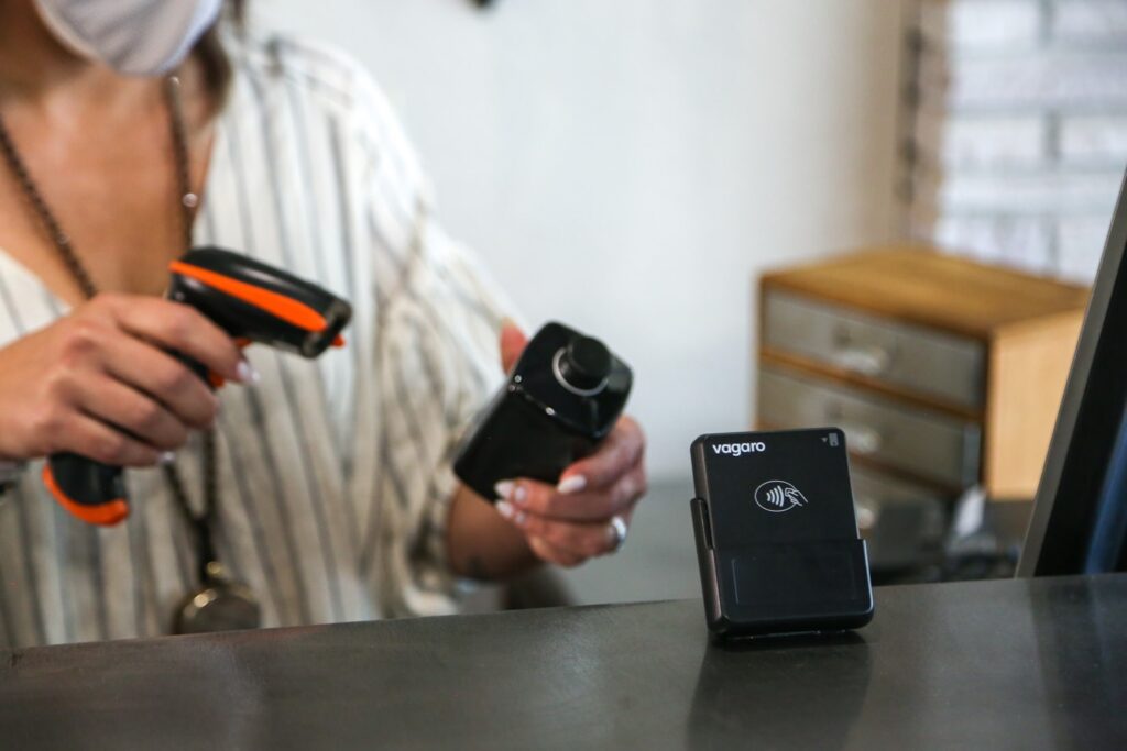 A woman at a front desk is scanning a hair styling product with a barcode scanner. The front desk has a credit card reader on it with the Vagaro logo on it.